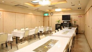 Un salón de conferencias con mesas largas, sillas, un reloj, un televisor y un jarrón decorativo.