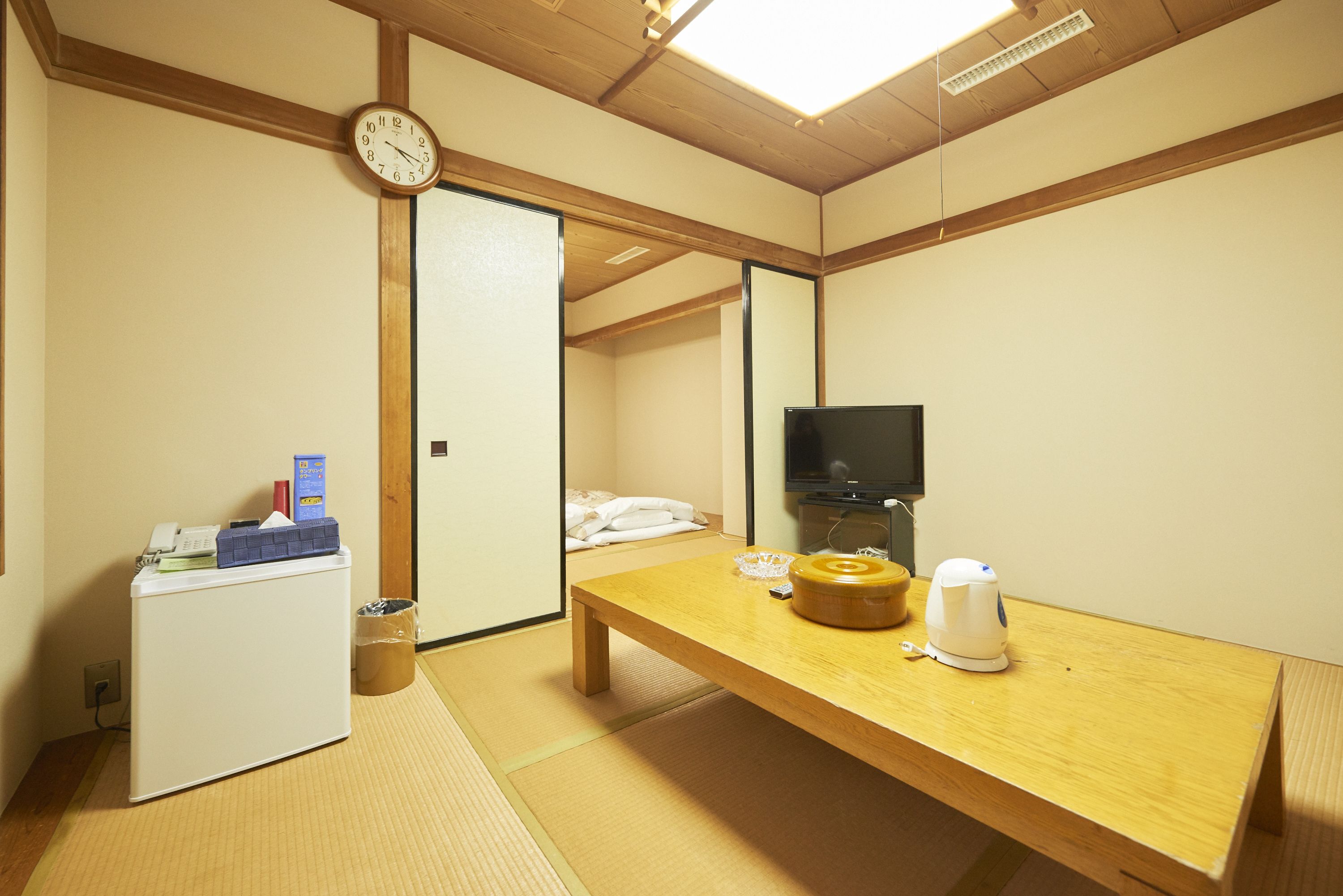 Ein Hotelzimmer mit Tatami-Böden, einem Holztisch, einem Fernseher, einem Kühlschrank und einer Wanduhr.