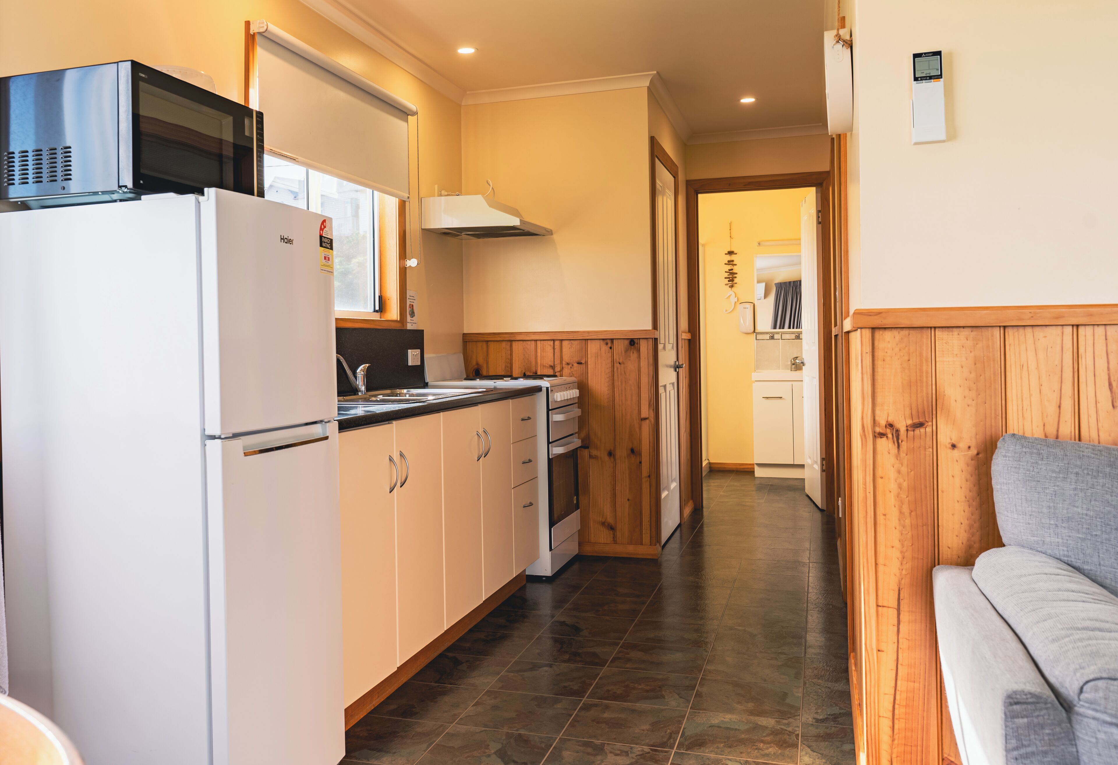 A compact kitchen with a refrigerator, microwave, and wooden cabinets. A grey sofa is visible in the adjacent living area.