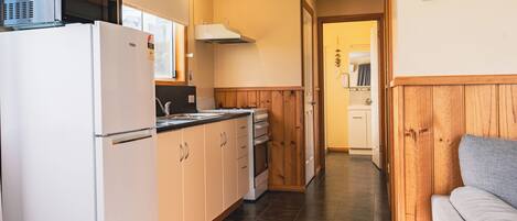 A compact kitchen with a refrigerator, microwave, and wooden cabinets. A grey sofa is visible in the adjacent living area.