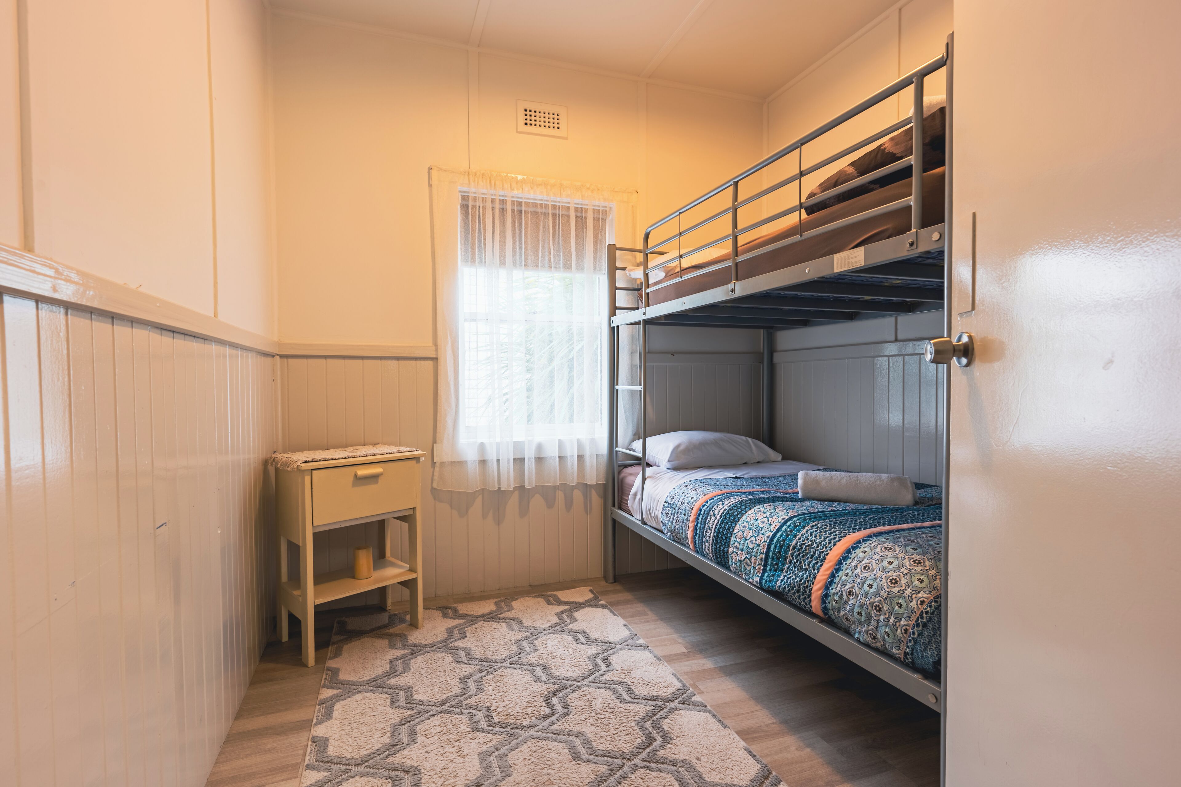 Habitación pequeña con literas, un escritorio y una ventana con cortinas traslúcidas.