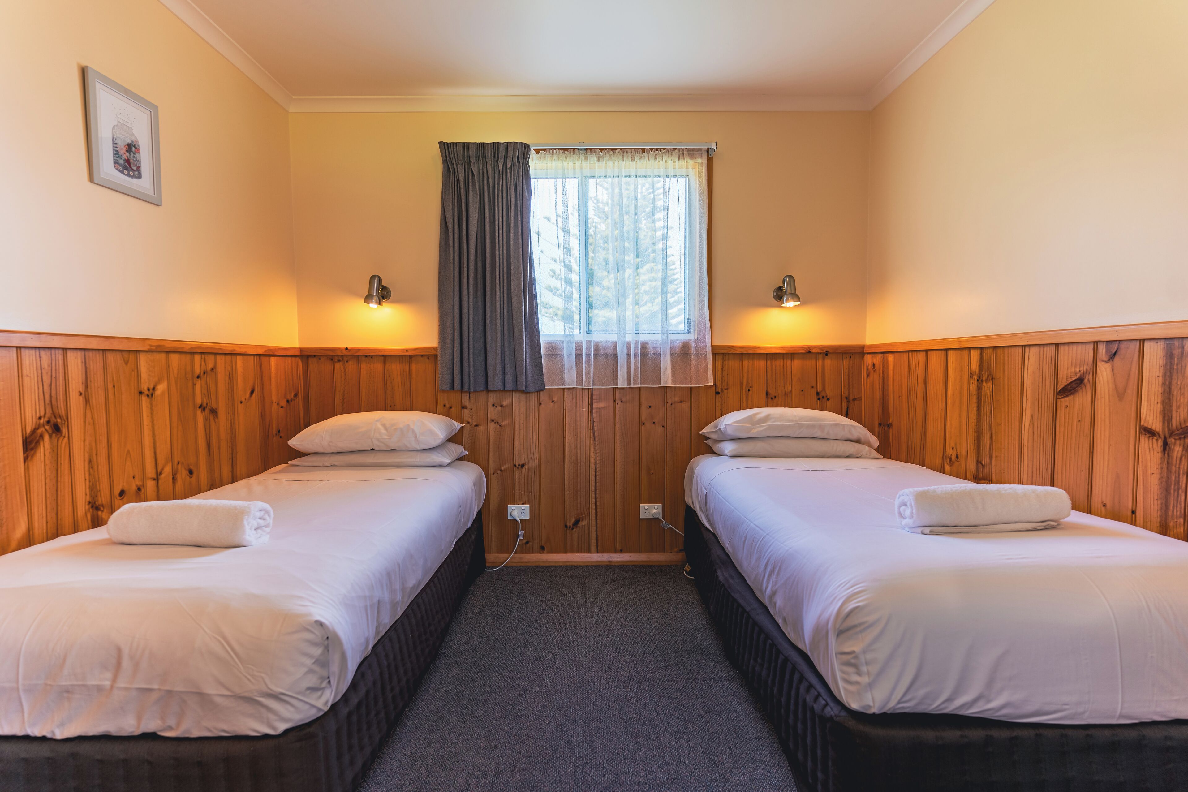 A room with two beds, wooden paneling, a window with a curtain, and wall-mounted lights.