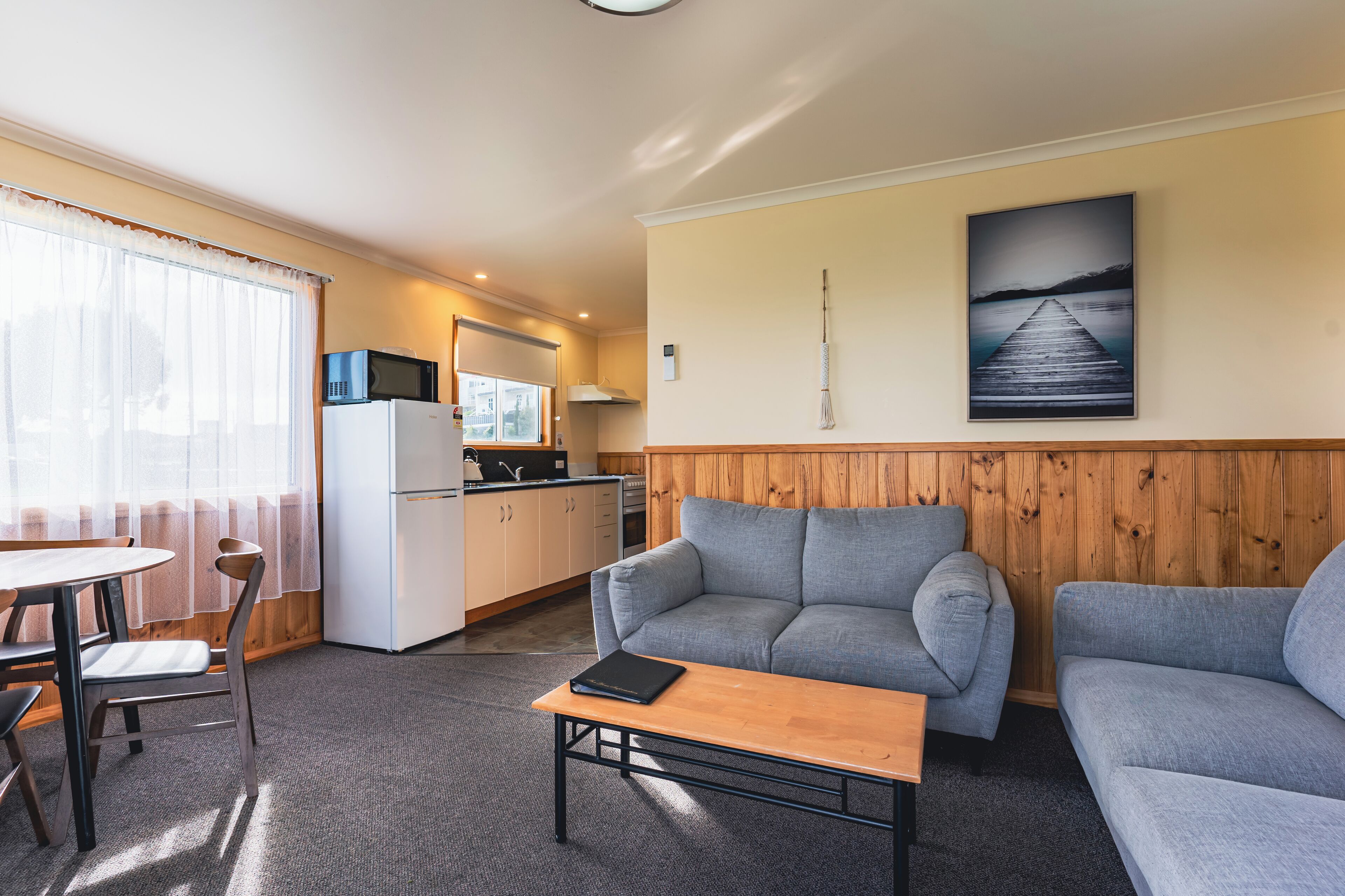 A compact living space with a kitchenette, a grey sofa, a dining table, and a painting of a pier on the wall.