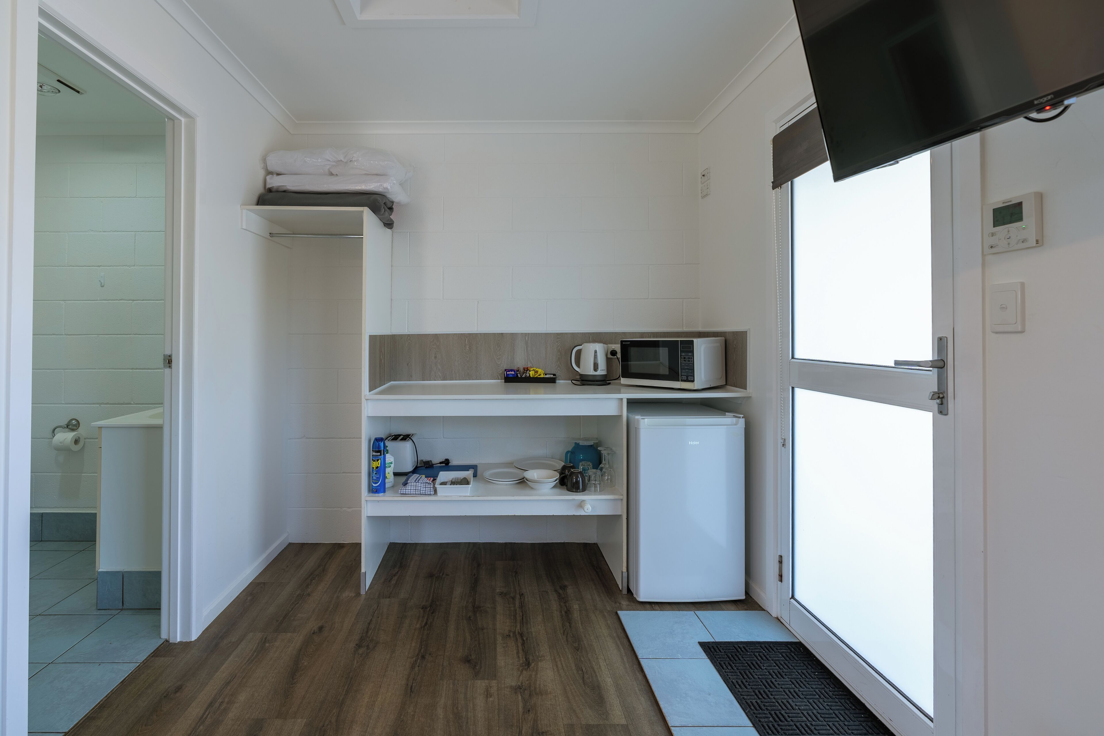 A compact kitchen with a microwave, kettle, and a small refrigerator. A white shelf is above the refrigerator, and a door with a window is on the right.