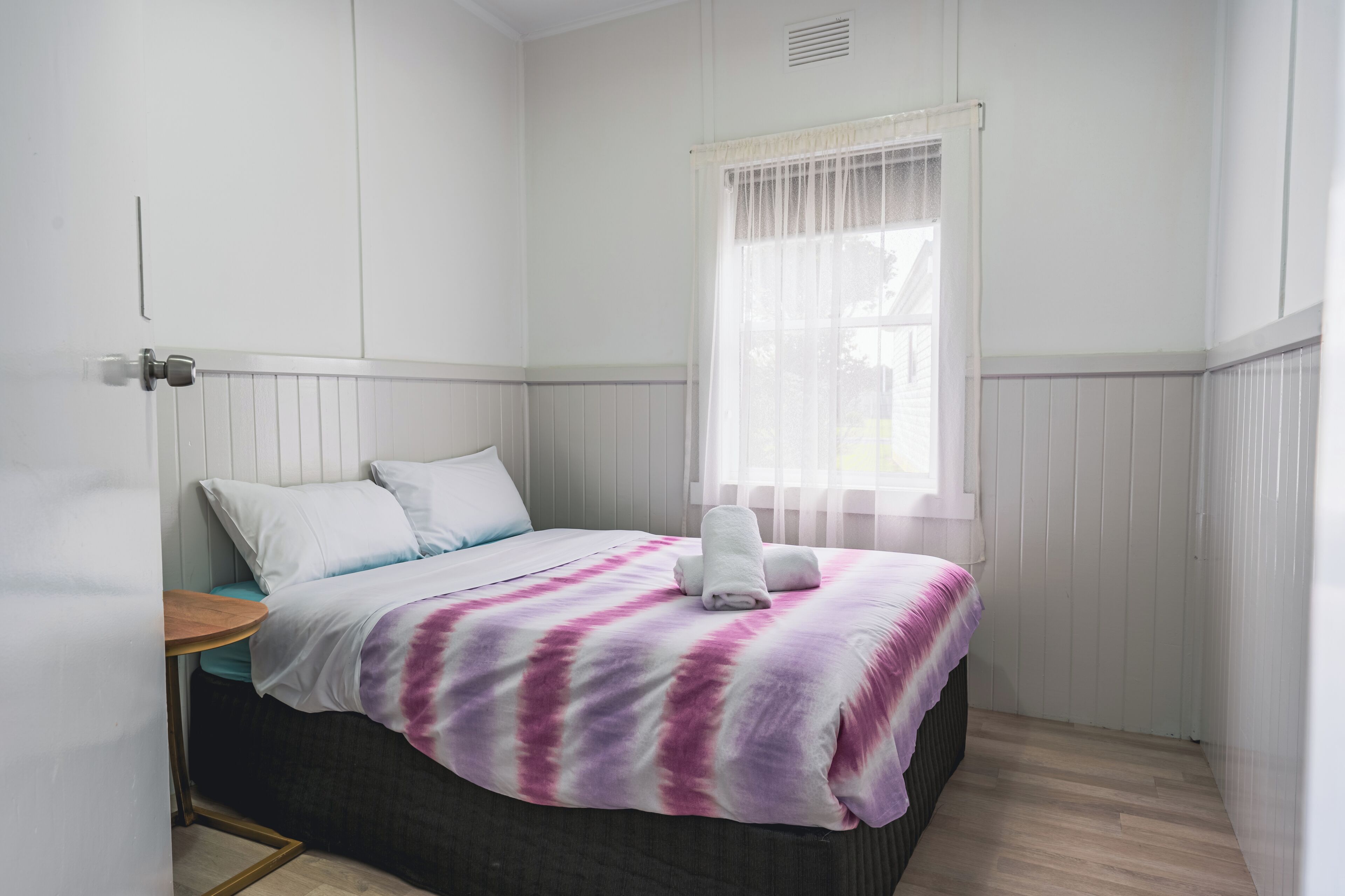 Una cama bien hecha con una manta morada y blanca, una mesita de noche pequeña y una ventana con cortinas transparentes.