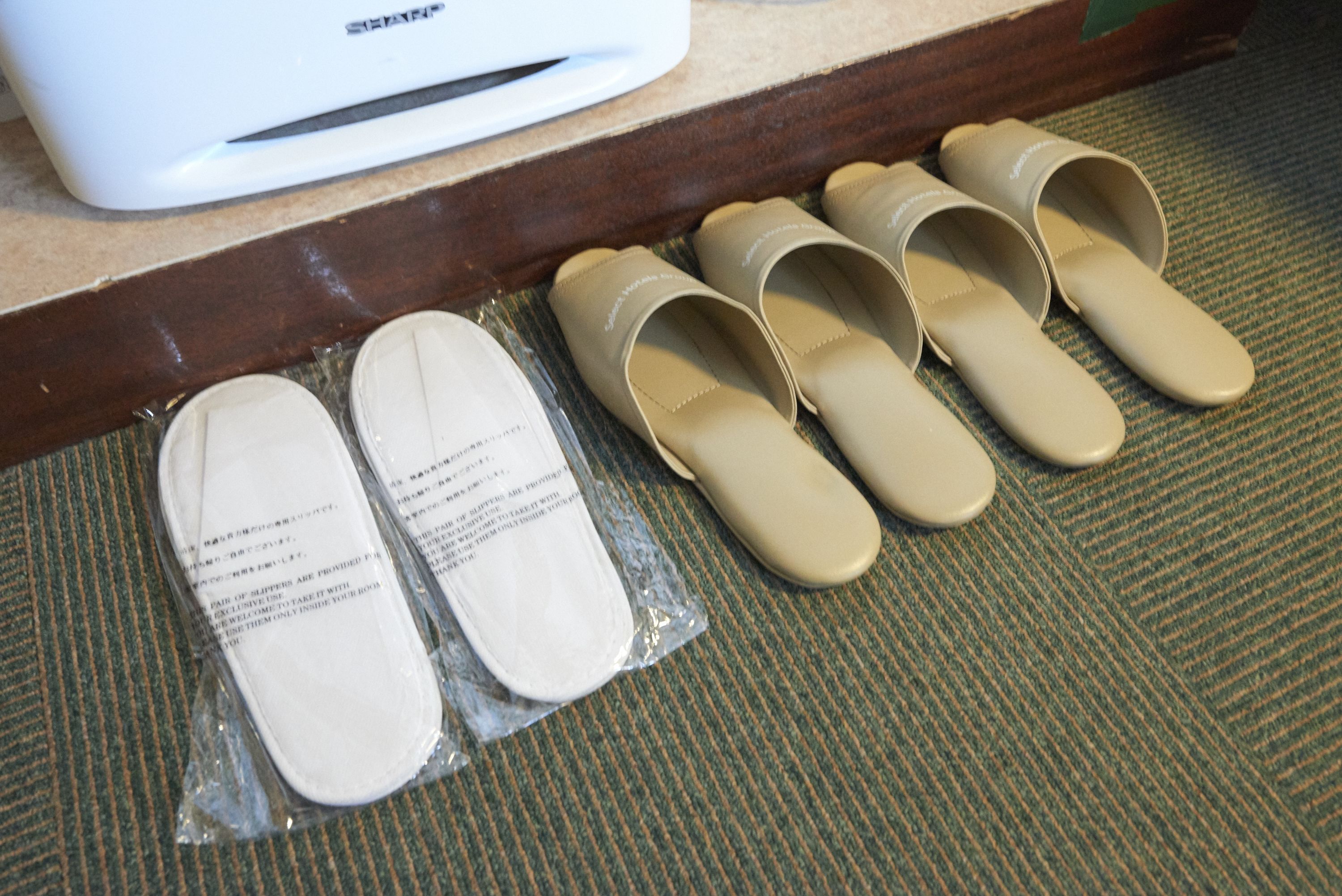 A pair of beige slippers and a pair of white slippers on a carpeted floor.