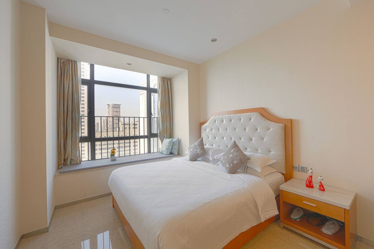 A hotel room with a large bed, a nightstand, a window with curtains, and a view of buildings outside.
