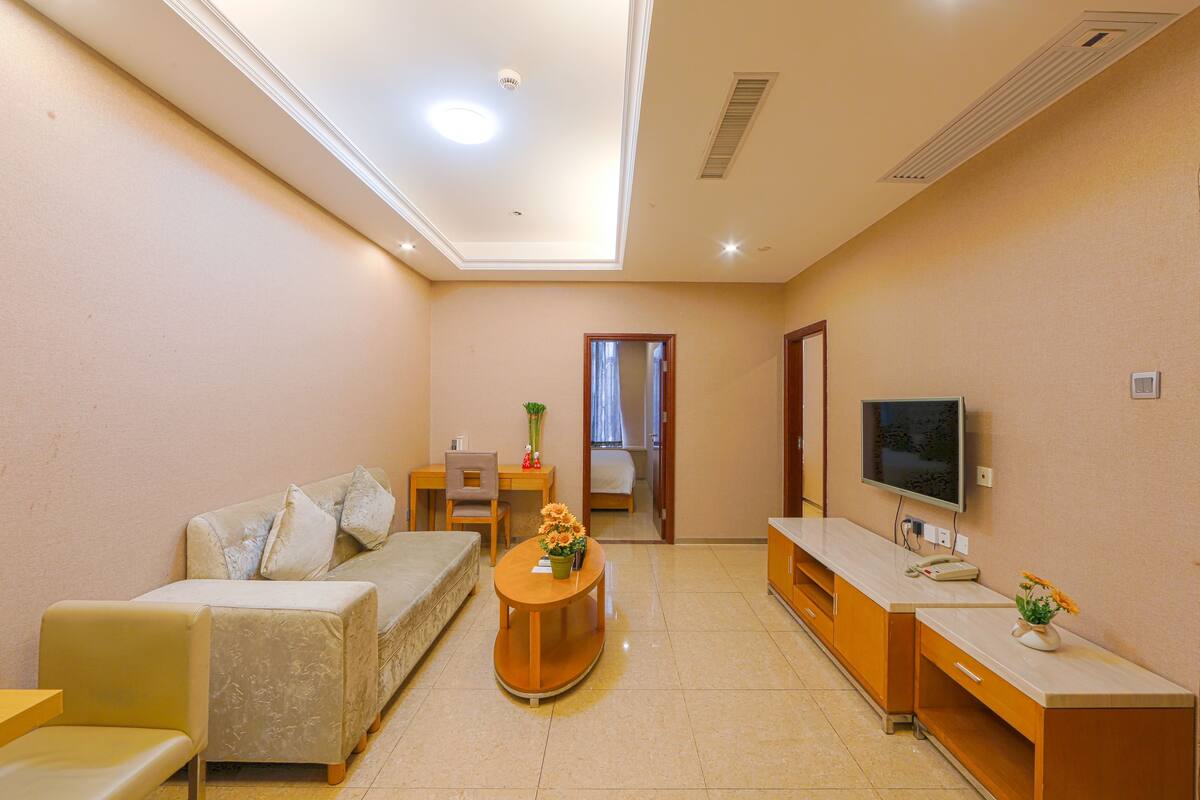 A modern hotel room with a sofa, a small table with flowers, a TV, and a desk with a chair.
