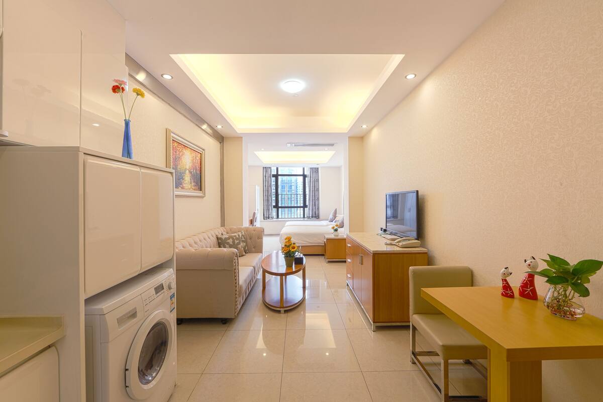 A modern hotel room with a washing machine, a sofa, a dining table, and a TV.