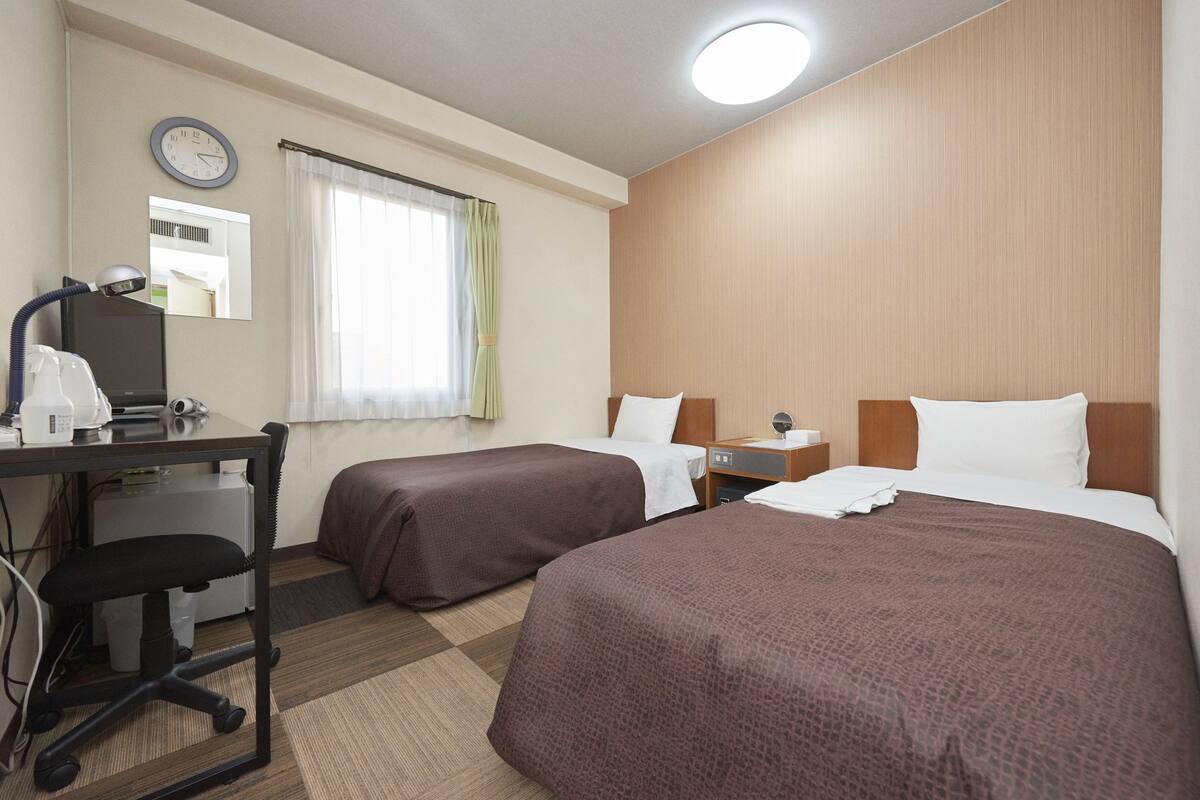 A hotel room with two beds, a desk with a chair, a desk lamp, a clock, and a window with curtains.