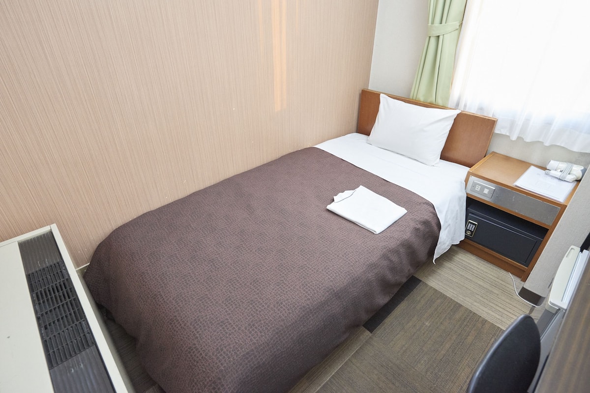 A hotel room with a single bed, a bedside table with a lamp, a small desk with a telephone, and a wall-mounted air conditioning unit.