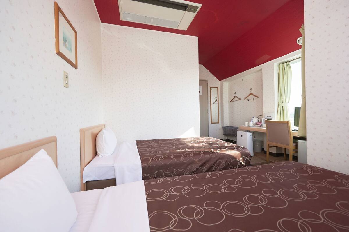 Quarto de hotel com duas camas, uma escrivaninha, uma cadeira e uma janela com cortinas.