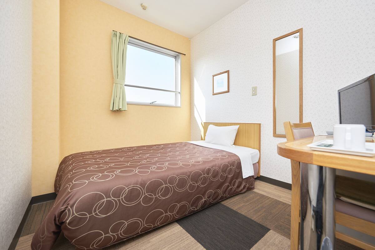 Quarto de hotel com cama, secretária e cadeira, janela com cortinas, espelho e um quadro na parede.