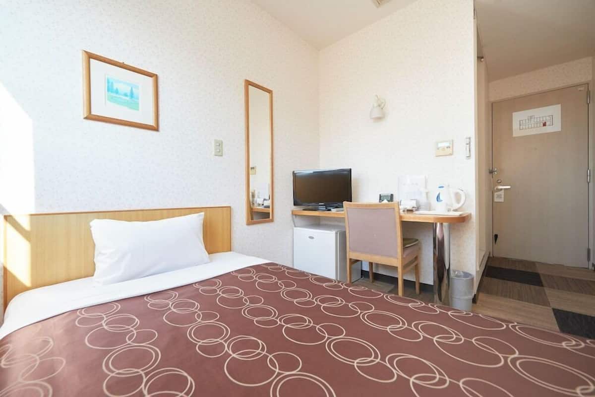 Quarto de hotel com cama, escrivaninha com computador, cadeira, televisão e pequeno espaço de cozinha.