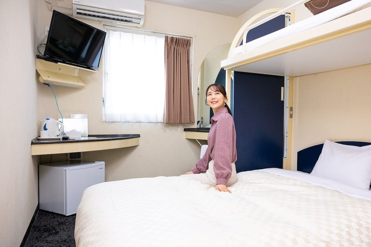 A hotel room with a bunk bed, a television on a stand, a small desk with a kettle, and a window with curtains.