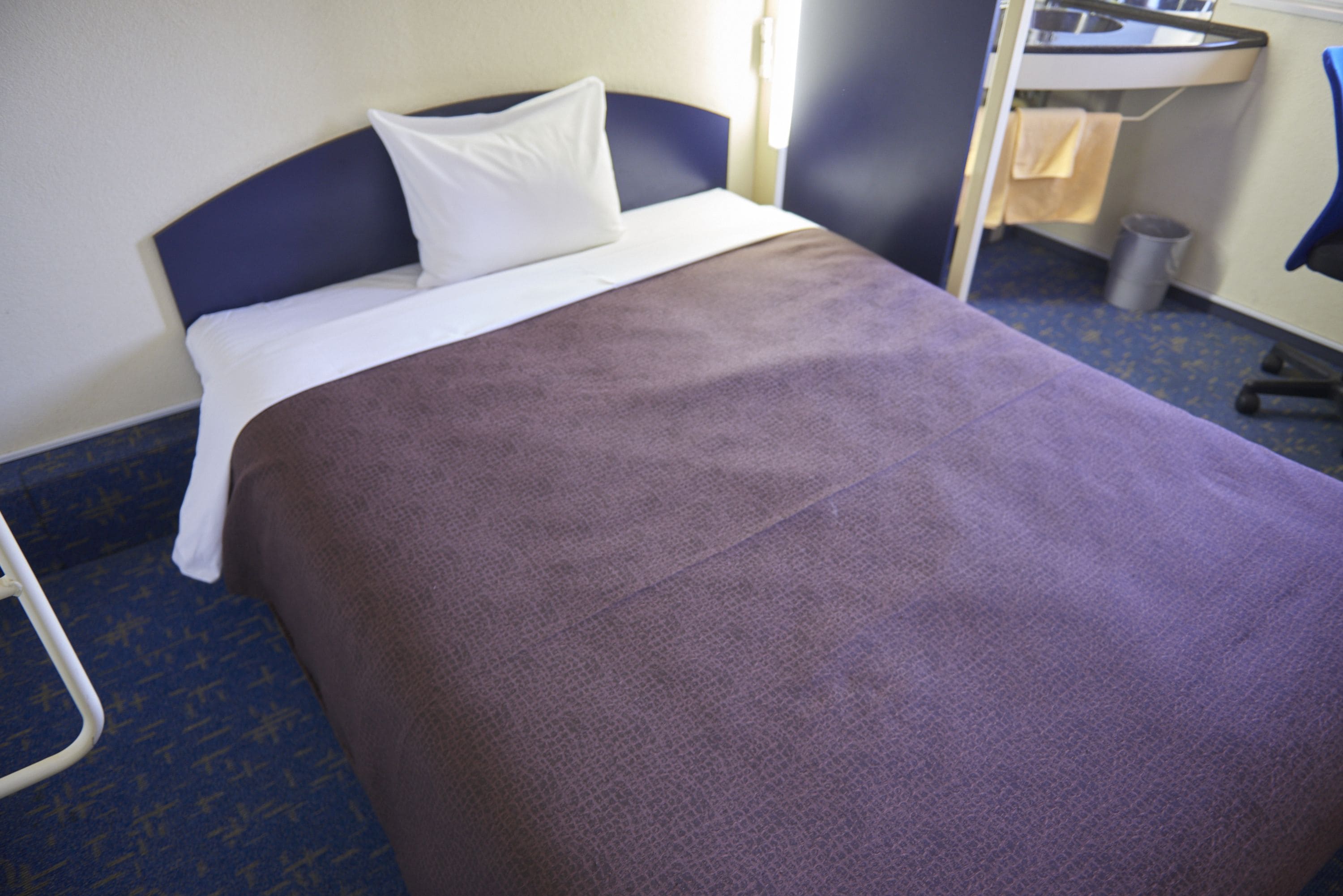 A hotel room with a bed, a blue patterned chair, a sink, and a trash bin.