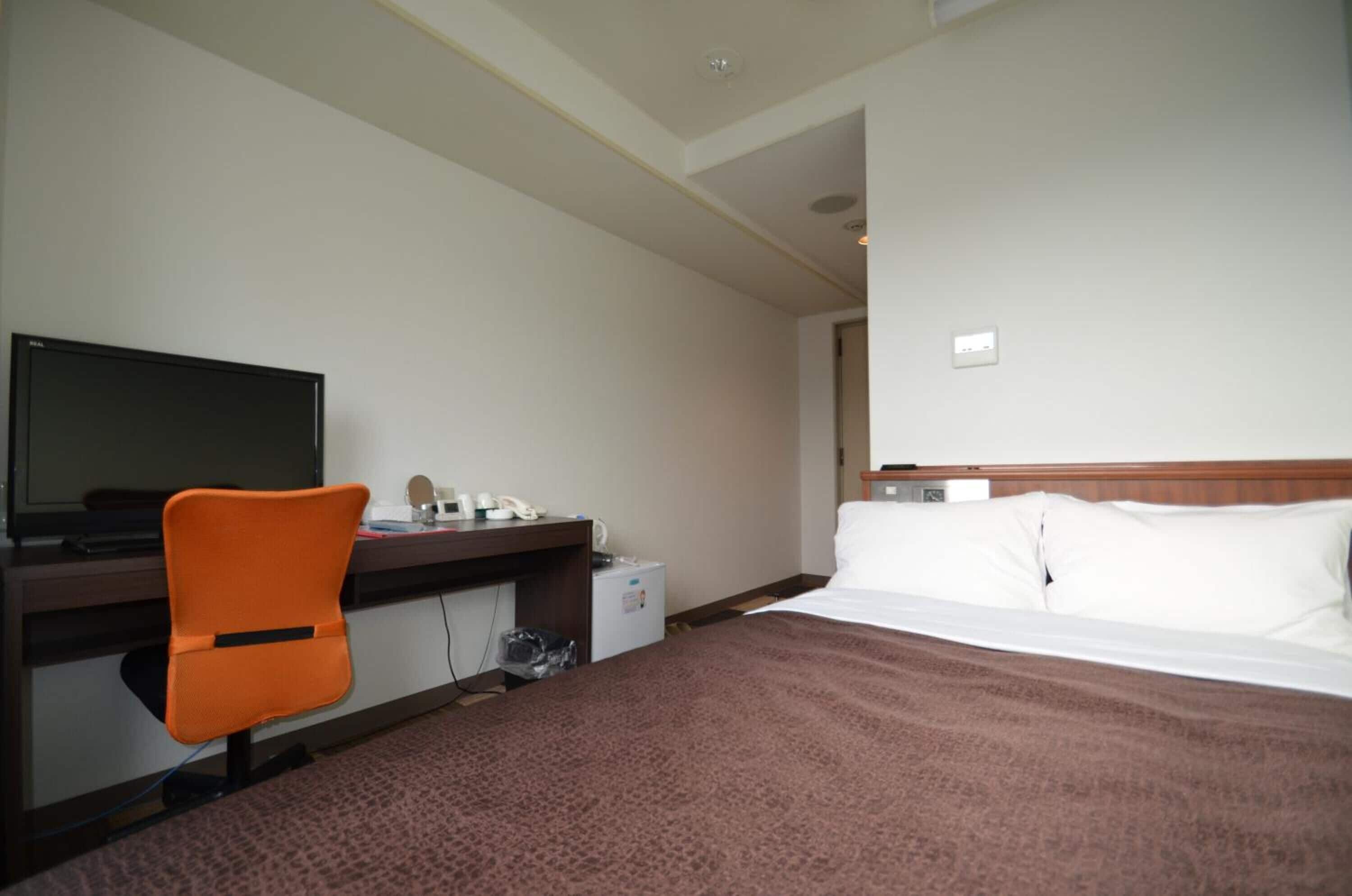 A hotel room with a bed, a desk with a computer, an orange chair, and a small refrigerator.