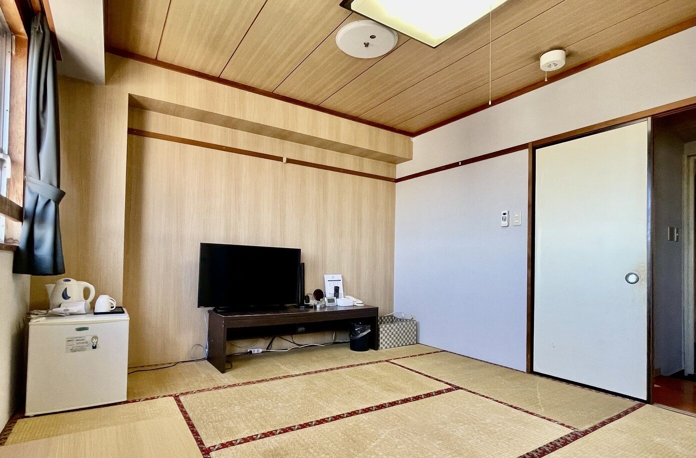 A room with a television on a wooden stand, a small refrigerator, a white wardrobe, and a tatami mat floor.