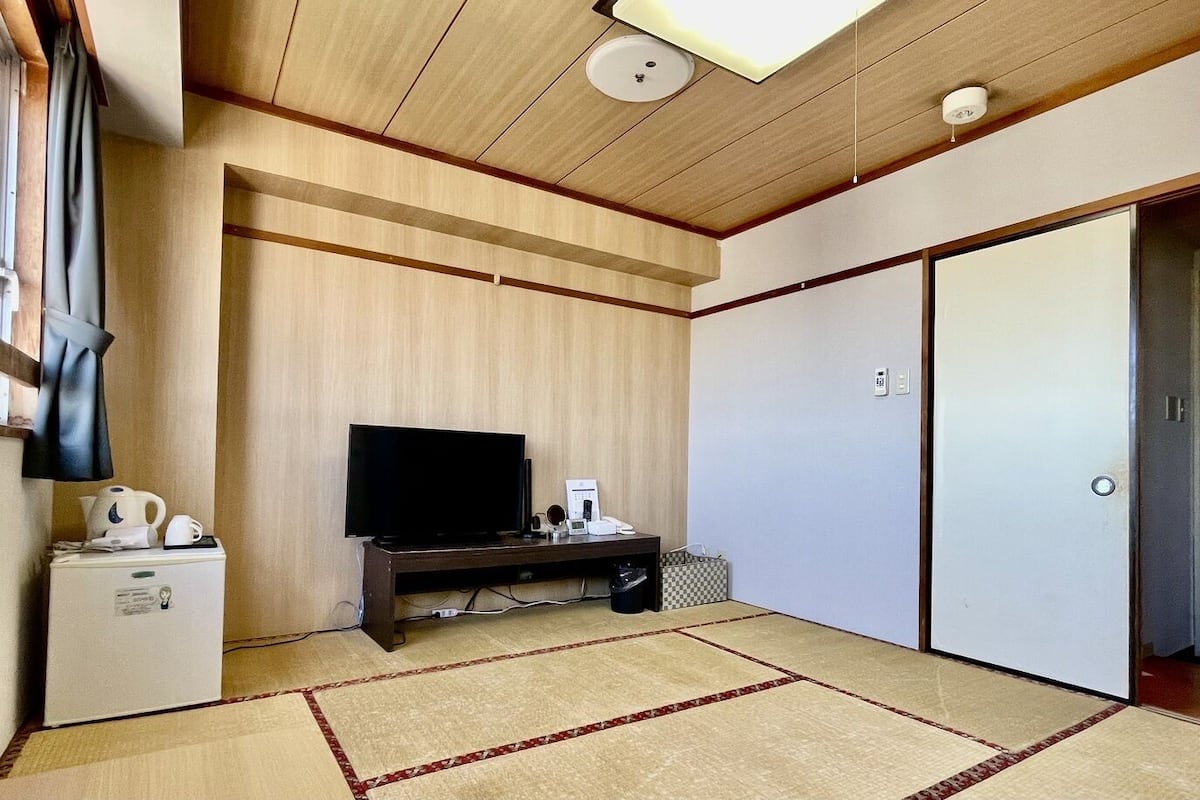 A room with a television on a wooden stand, a small refrigerator, a white wardrobe, and a tatami mat floor.