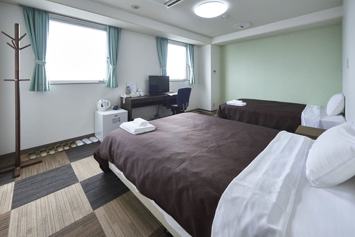 A hotel room with a bed, a desk, a chair, a coat rack, a small refrigerator, and a window with curtains.