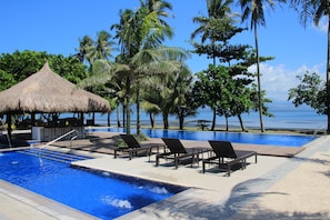 Zona de piscina con sillones reclinables y una estructura de techo de paja, rodeada de palmeras y vista al mar.