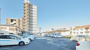 A parking lot with several cars, including a white car with "Nakamura-Tome" on the side, and a tall building with multiple balconies in the background. - Hotel Select Inn Isehara (Isehara)