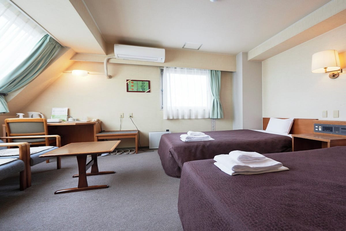 A hotel room with two beds, a desk, a chair, a small table, and a window with curtains.