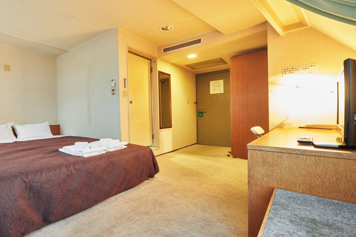 A hotel room with a large bed, a desk with a lamp, a mirror, and a door leading to another room.
