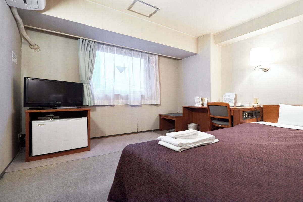 A hotel room with a bed, a desk, a chair, a television, a mini-fridge, and a window with curtains.