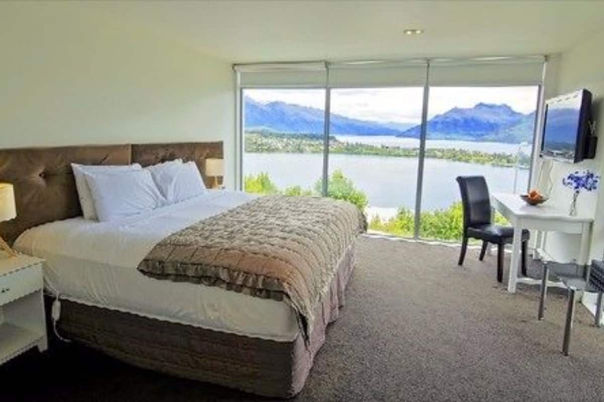A bedroom with a bed, a desk, a chair, and a large window offering a view of a lake and mountains.