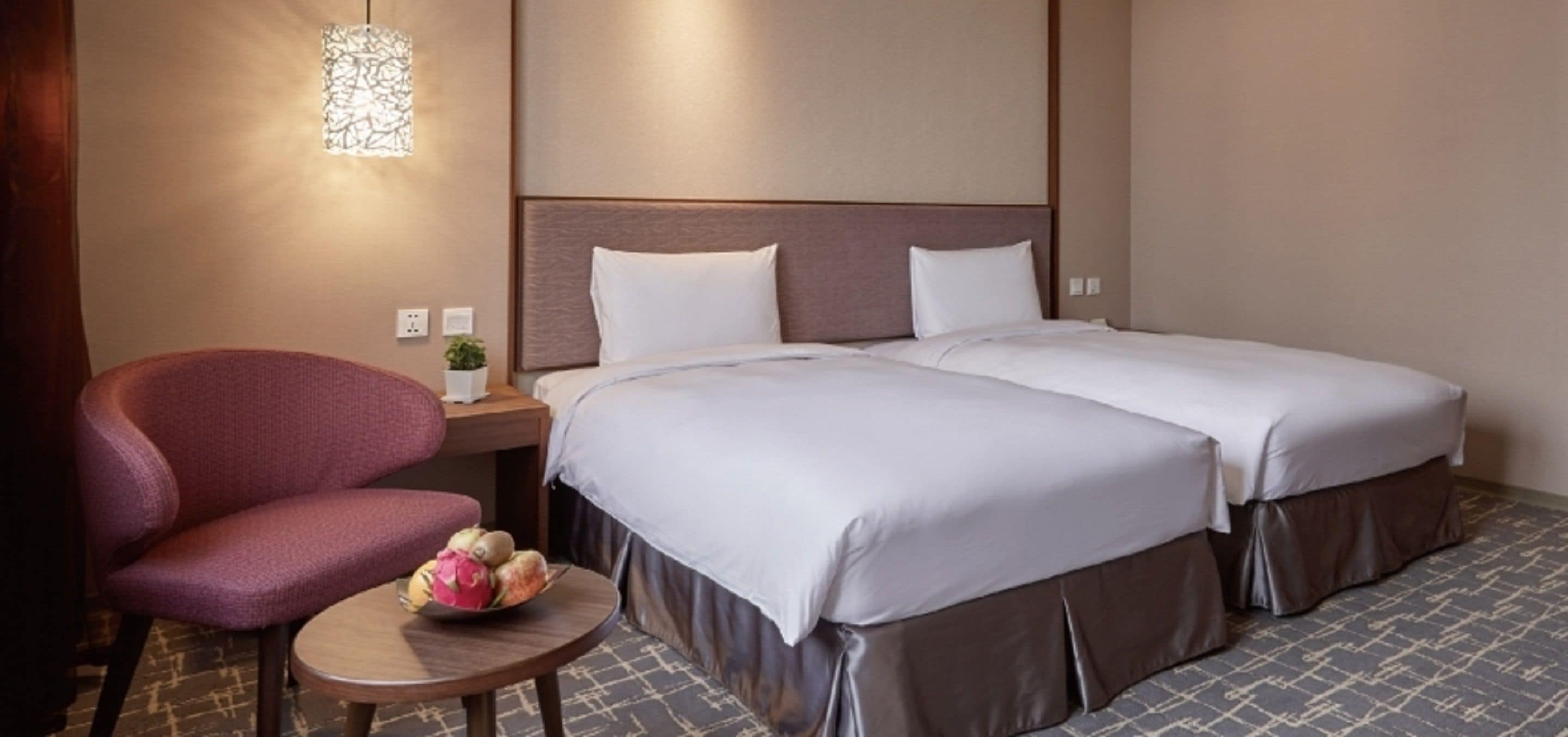 Habitación de hotel con cama, una silla, una pequeña mesa con frutero y una lámpara fijada a la pared.