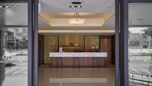 A reception area with a modern design, featuring a reception desk, three bar stools, a chandelier, and a large window offering a view of the street.