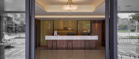 A reception area with a modern design, featuring a reception desk, three bar stools, a chandelier, and a large window offering a view of the street.