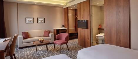 A hotel room with a sofa, armchair, TV, and a small table with a bowl of fruit.