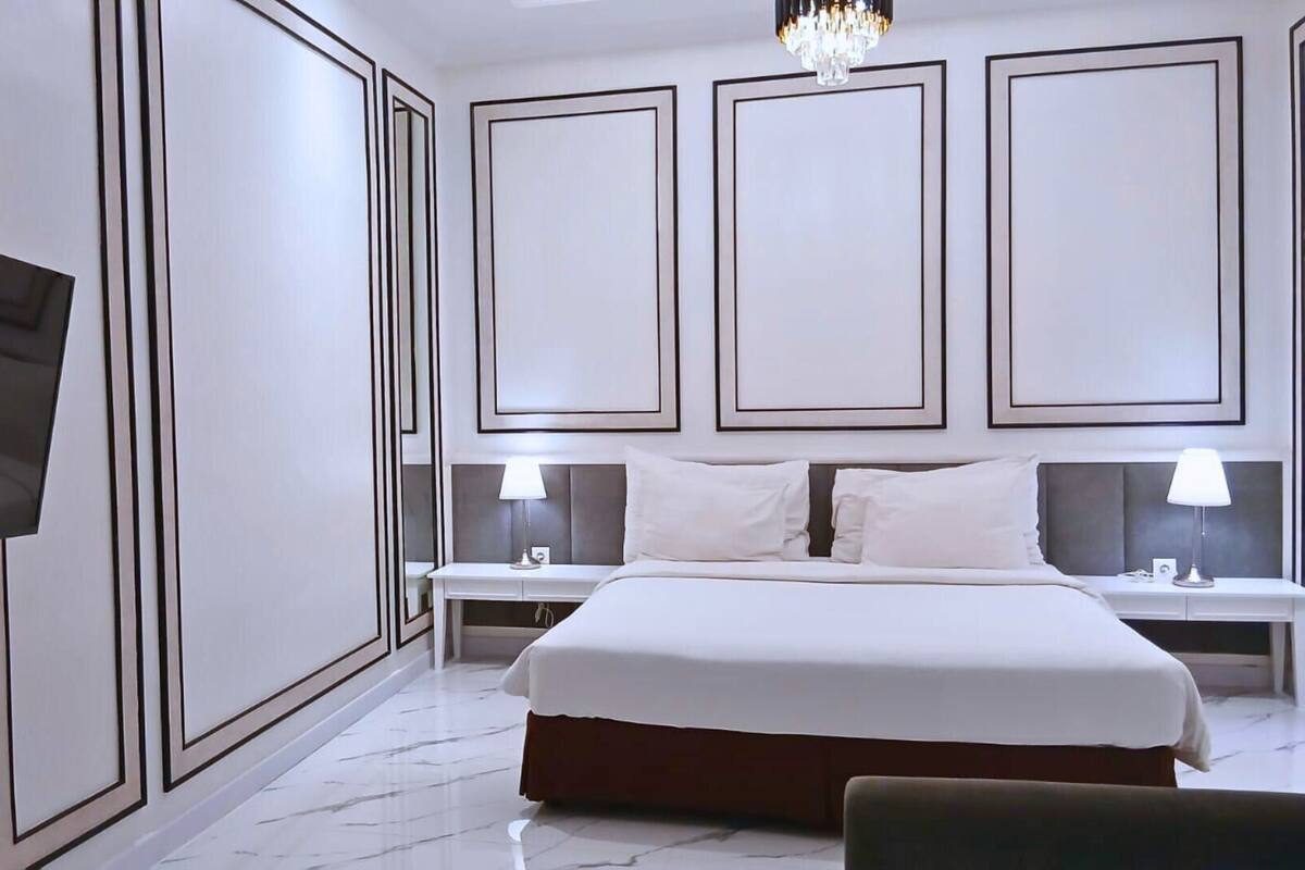 Une chambre d’hôtel moderne avec un grand lit, deux tables de chevet avec des lampes, un téléviseur à écran plat fixé au mur et un lustre.