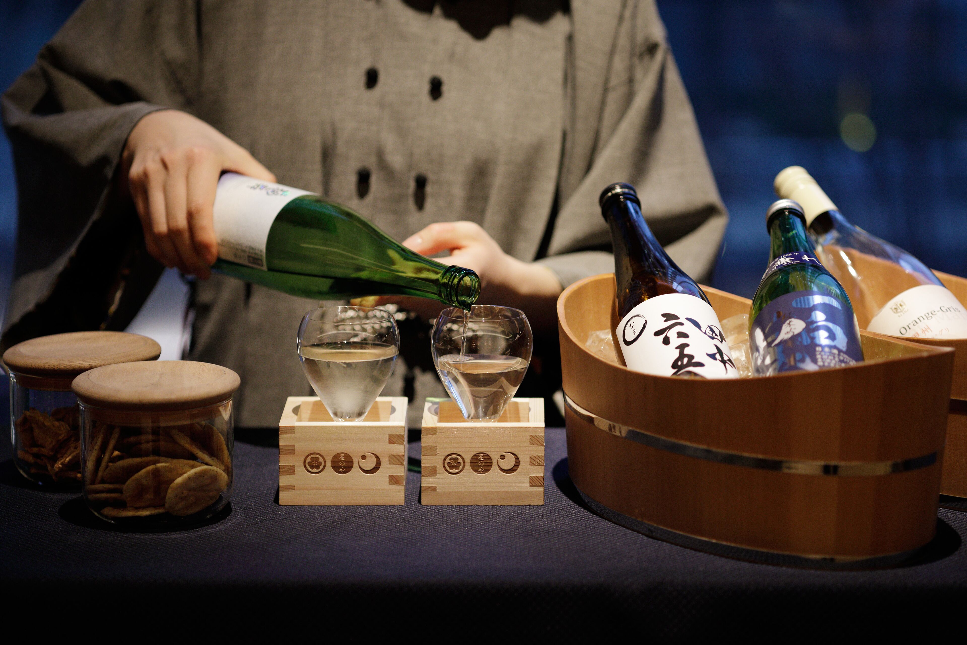 A person pouring a drink from a green bottle into a glass. There are wooden blocks with symbols, a wooden bowl, and bottles in the background.