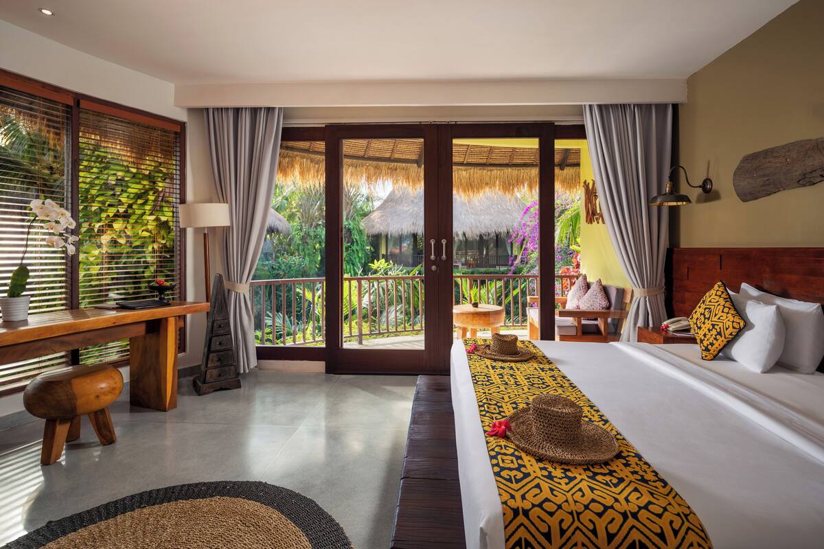 A hotel room with a large bed, a wooden desk, a small table, and a balcony with a thatched roof.