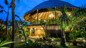 Un edificio grande de forma abovedada con techo de paja, rodeado de exuberante vegetación tropical e iluminado por una cálida luz interior.