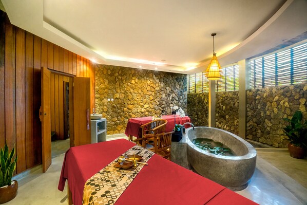 Un cuarto de spa con pared de piedra, una puerta de madera, una mesa roja con una toalla, una bañera de piedra y una lámpara colgante.