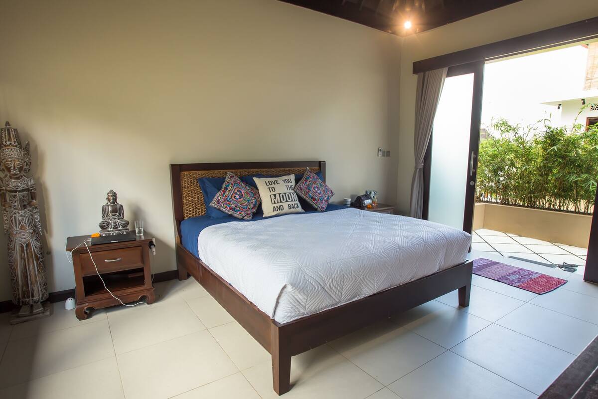 A bedroom with a bed, a nightstand, a decorative statue, and a view of a balcony with plants.