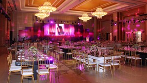 Un salón de banquetes con largas mesas dispuestas para un evento formal, candelabros y un escenario con una gran pantalla.