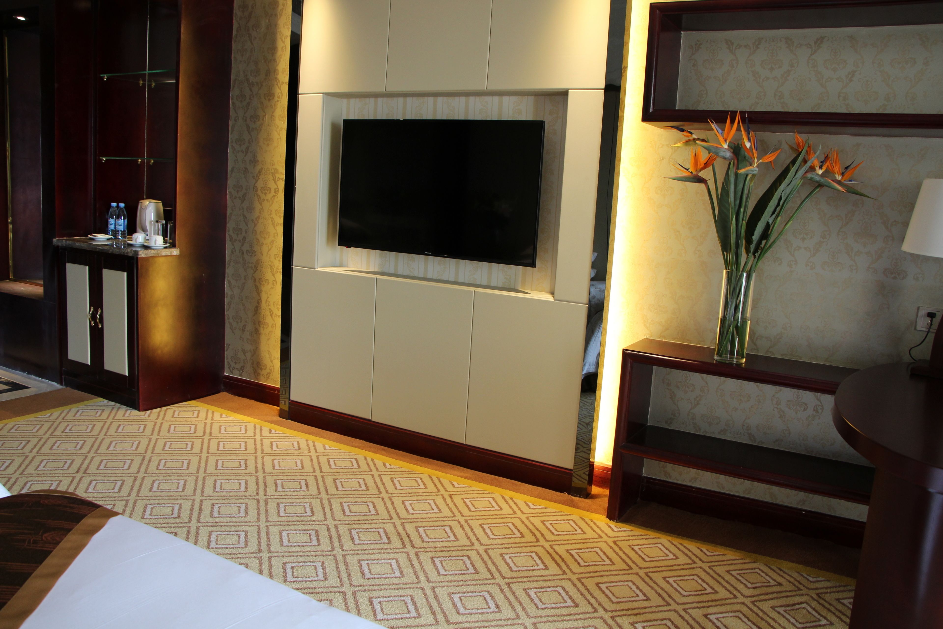 A hotel room with a flat-screen TV mounted on the wall, a coffee machine, a vase with flowers, and a patterned carpet.