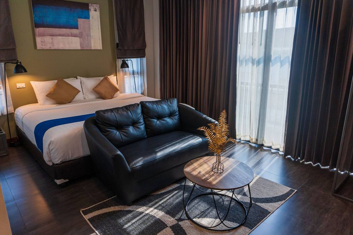 A hotel room with a bed, a black leather sofa, a small round table, and a decorative plant.