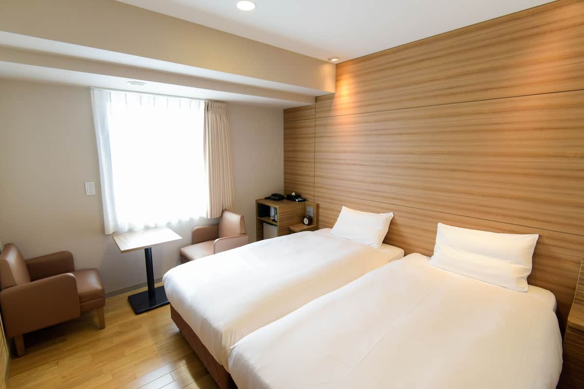 Quarto de hotel com duas camas, uma escrivaninha, uma cadeira e uma janela com cortinas.