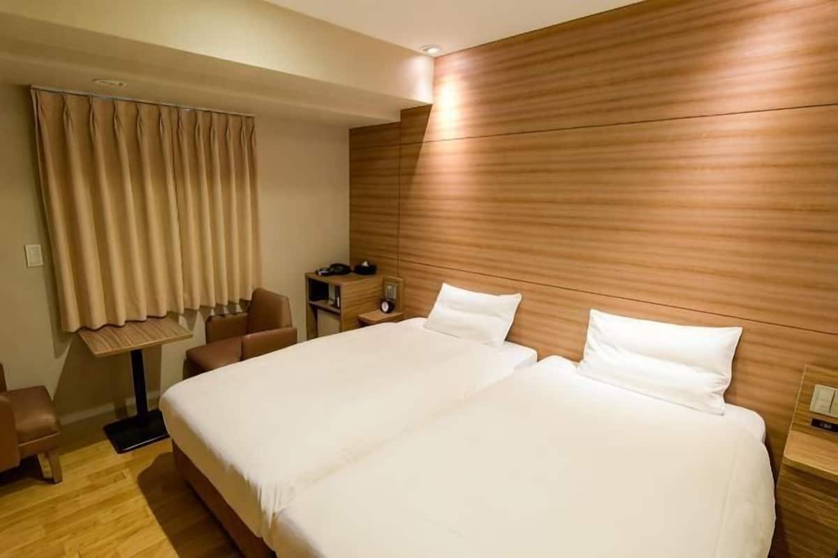 Cama de casal com lençóis brancos, cabeceira de madeira, escrivaninha com cadeira e janela com cortinas.