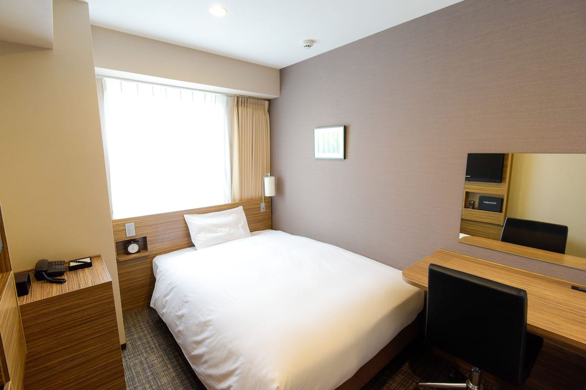 Quarto de hotel com cama, escrivaninha, cadeira, telefone, relógio e janela com cortinas.