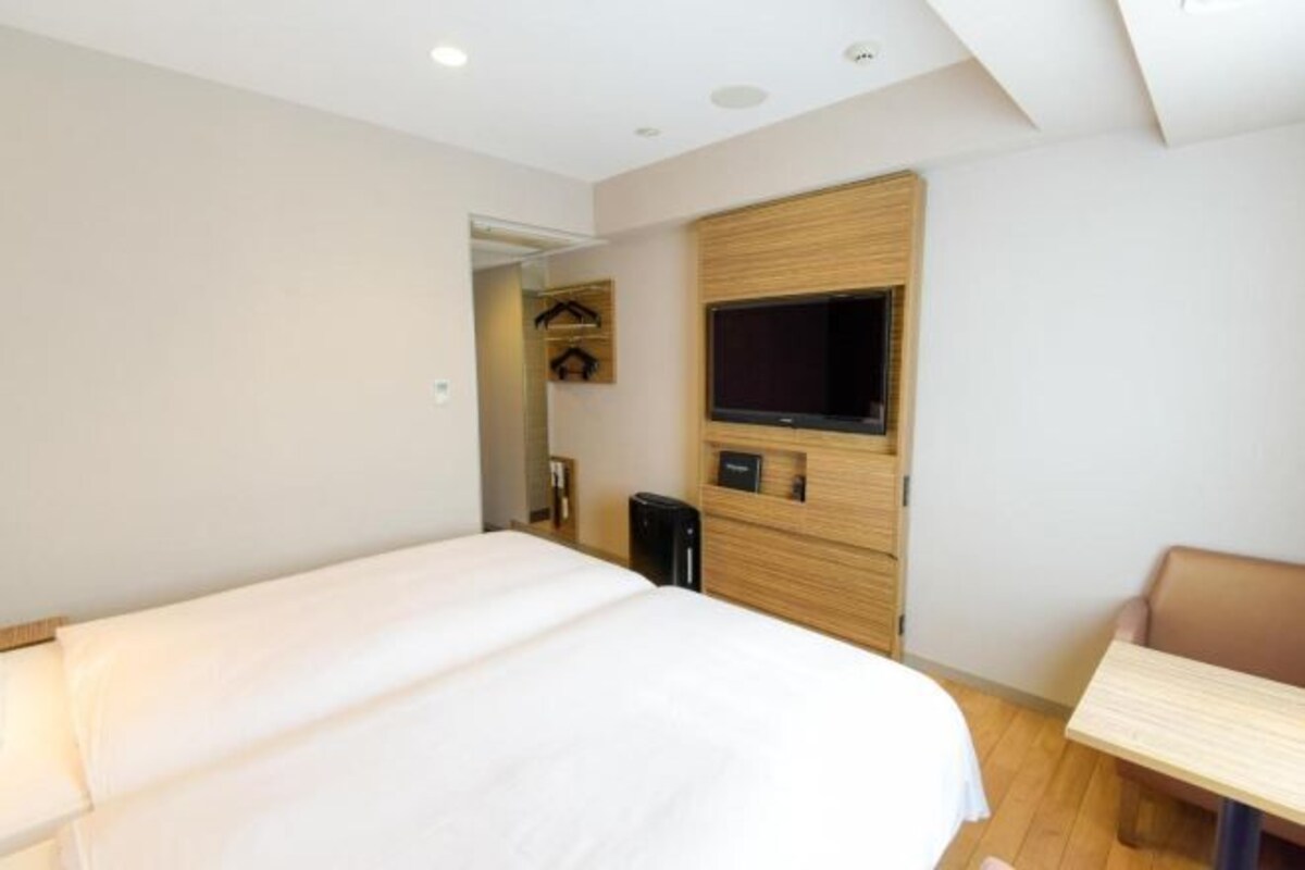 A hotel room with a bed, a TV mounted on the wall, a small table, and a closet with hanging clothes.