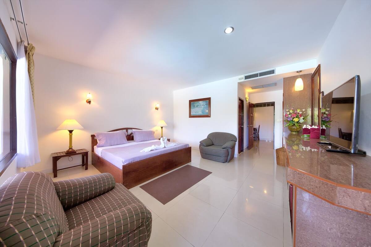 Habitación de hotel con una cama, dos lámparas de noche, una silla, un sofá, un baño con espejo y un florero.