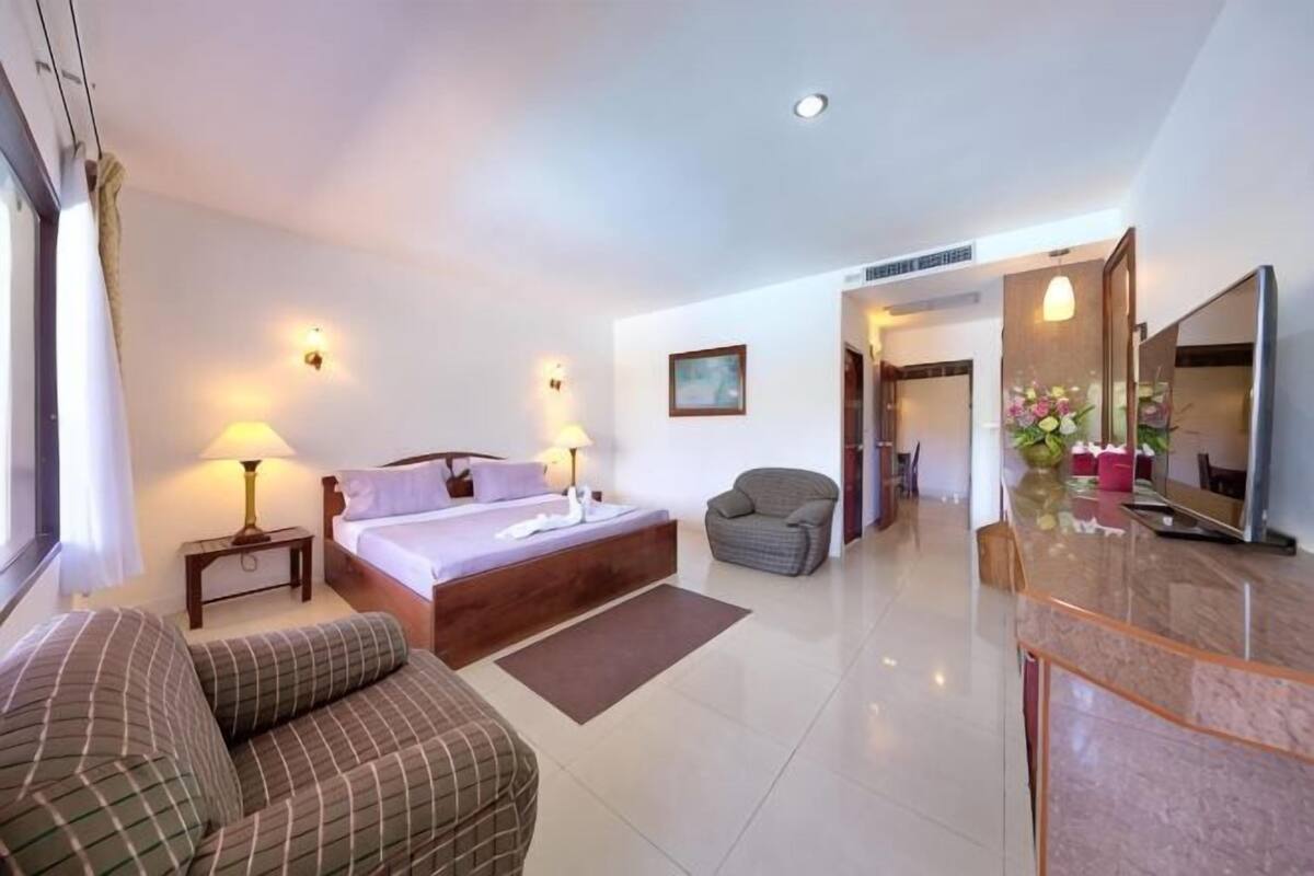 Habitación de hotel con cama, dos mesitas de noche con lámparas, un sofá, un escritorio con espejo, y un baño visible a través de una puerta abierta.