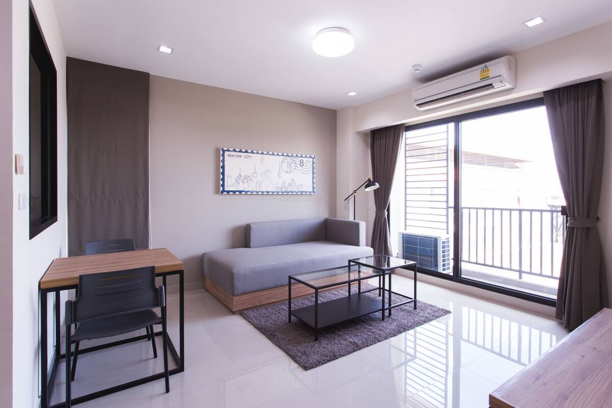 Una sala de estar moderna con sofá, mesa de comedor y mesa de centro. Hay un ventanal con cortinas, un equipo de aire acondicionado y un cuadro en la pared.